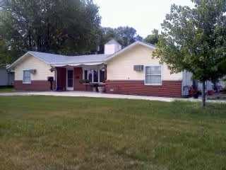 Single-story residential building with light yellow siding and red brick lower exterior, surrounded by green grass and trees under a clear sky.