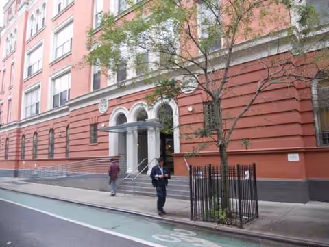 Red brick building with arched entrance, steps, and pedestrians along a city sidewalk.