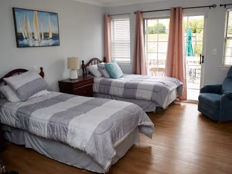 A bright bedroom with two twin beds featuring gray and white striped bedding, a wooden nightstand with a lamp between the beds, a blue armchair, and a sliding glass door leading to an outdoor patio. The room has light-colored walls, wooden flooring, and a painting of sailboats on the wall.