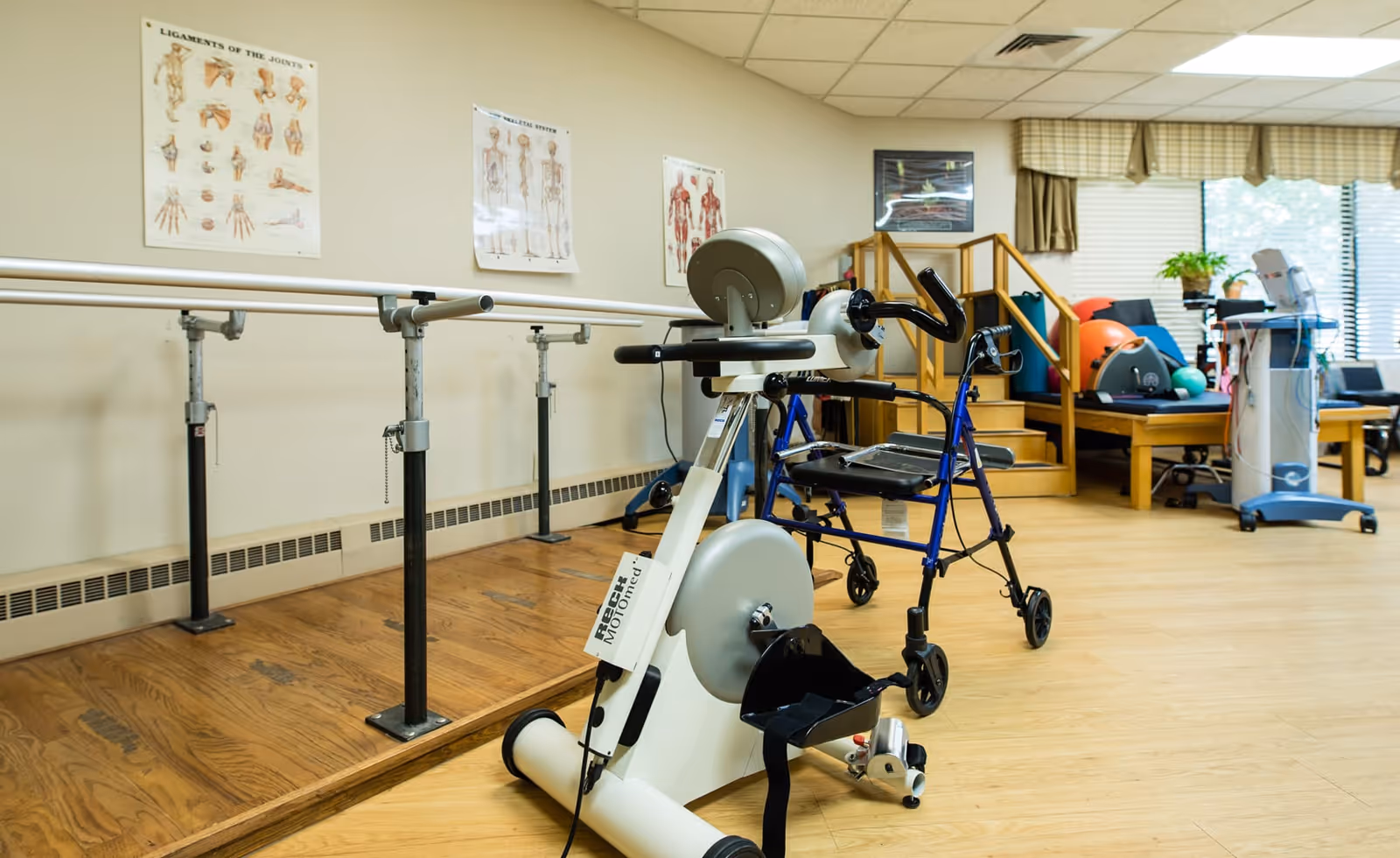Physical therapy room with exercise equipment including a stationary bike, walker, parallel bars for walking support, and various therapy tools. Anatomical posters are displayed on the wall, and there is a window with blinds and a valance letting in natural light.