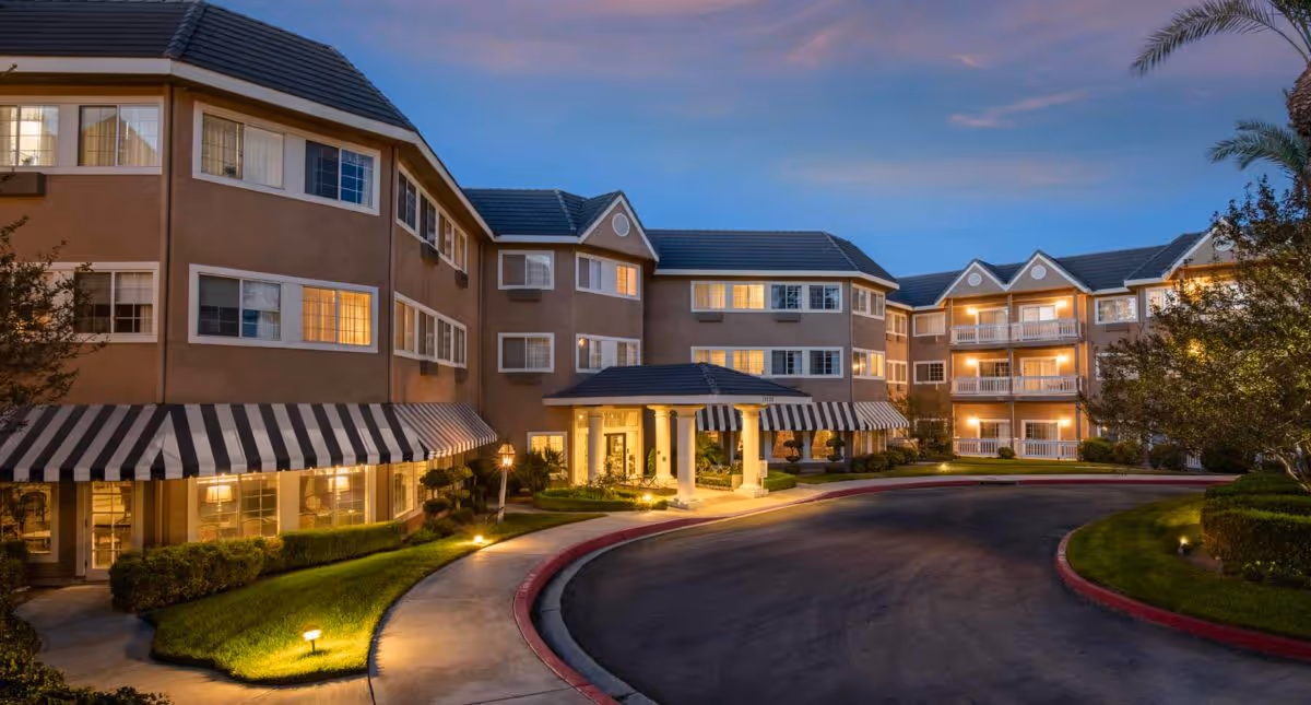 Exterior view of a multi-story assisted living community building at dusk with warm lights glowing from the windows. The building features striped awnings over the ground floor windows and a covered entrance with columns. The driveway curves in front of the entrance, surrounded by well-maintained landscaping and trees.