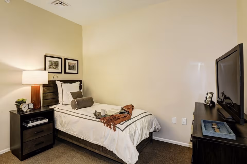 A cozy bedroom with a single bed featuring white bedding and decorative pillows. Next to the bed is a dark wooden nightstand with a lamp, a small plant, and two clocks. Above the bed are two framed pictures. Across from the bed is a dark wooden dresser with a flat-screen TV and a framed photo on top. The walls are painted a light beige color.