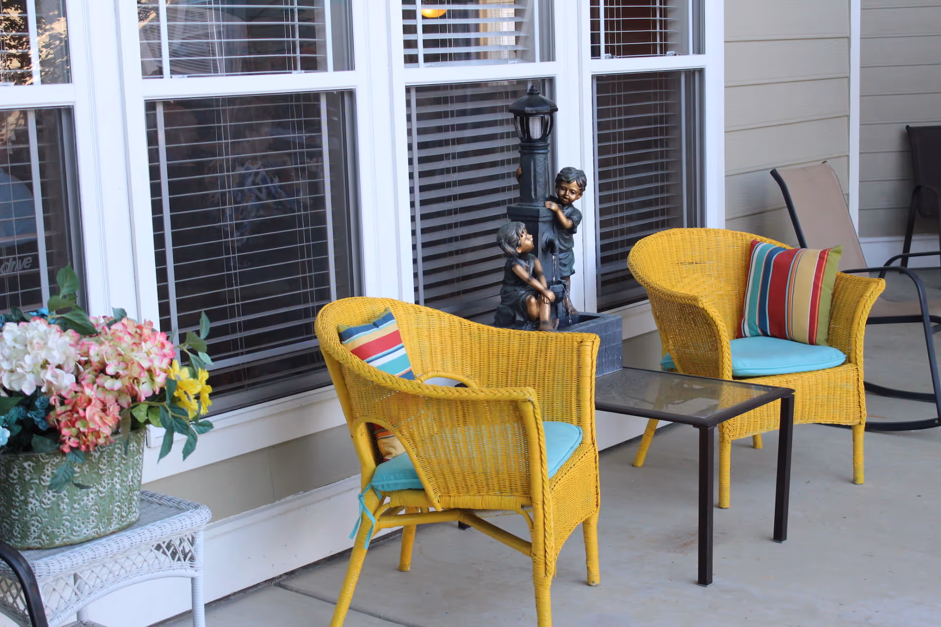 A porch seating area with two yellow wicker chairs, a small glass-top table, potted flowers and a decorative statue in front of large windows.