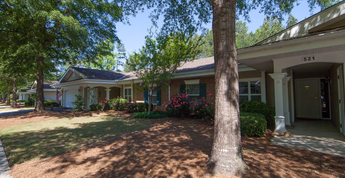 Exterior view of a single-story brick building with a covered entrance and a garage. The building is surrounded by trees, bushes, and flowering plants, with a well-maintained lawn and shaded areas. The address number 521 is visible above the entrance.
