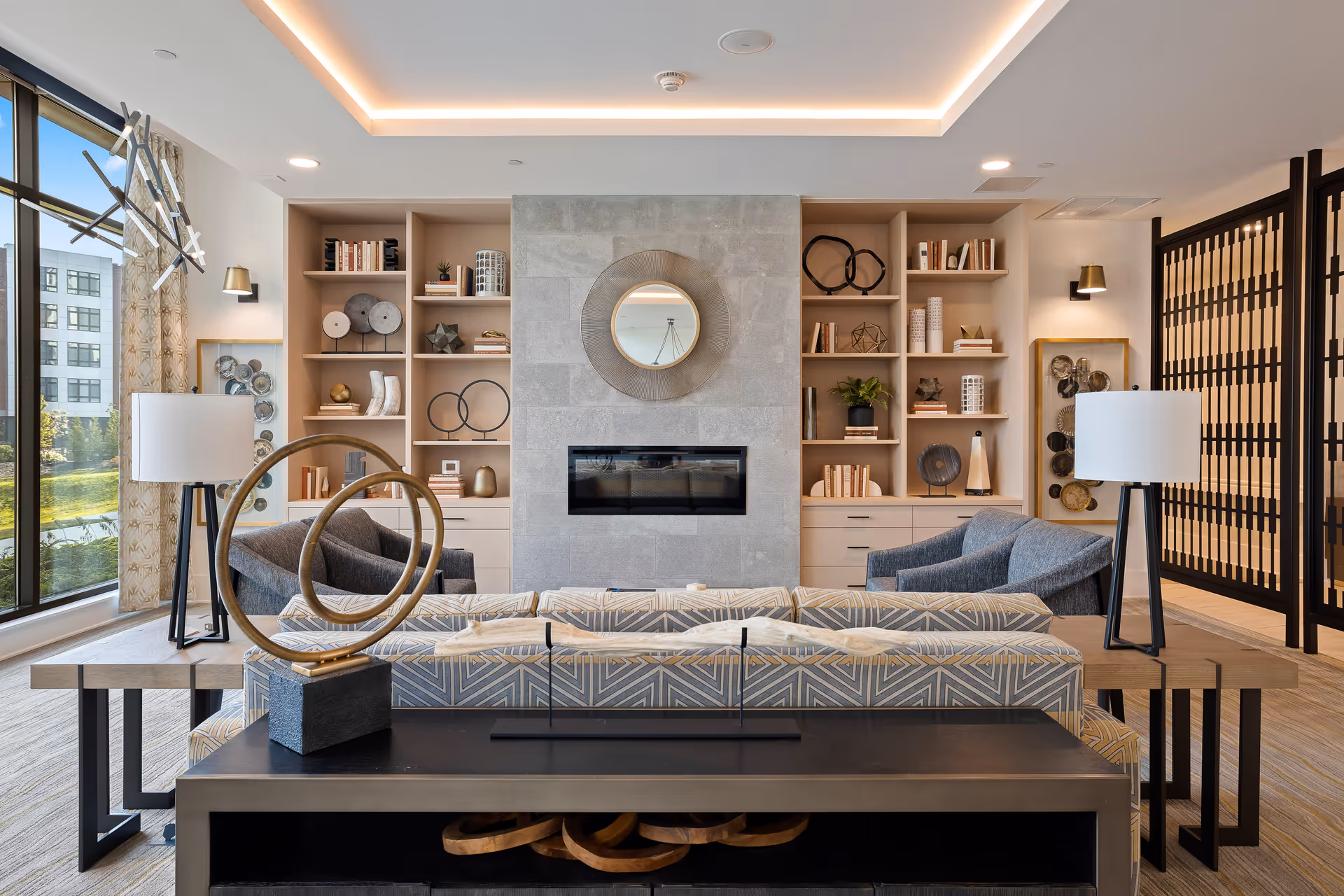Modern living room with a patterned sofa, two gray armchairs, two side tables with white lamps, built-in wooden shelves filled with decorative items and books, a stone fireplace with a round mirror above it, and large windows letting in natural light.