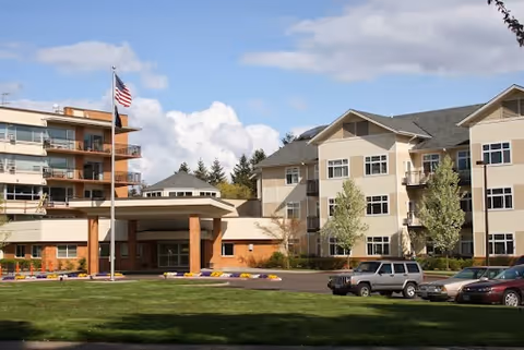 Front exterior of a multi-story retirement community building with a flagpole, lawn and parked cars.