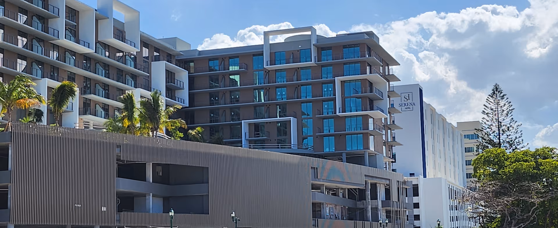 Exterior view of a multi-story senior living building with balconies, palm trees, and a parking structure under a blue sky.