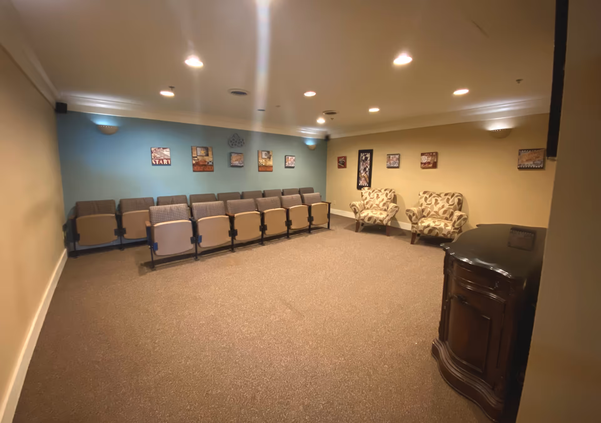 A small theater or presentation room with two rows of beige and brown cushioned chairs facing a blank wall. The room has beige and blue walls decorated with framed pictures and two patterned armchairs placed against the right wall. The floor is carpeted, and the ceiling has recessed lighting.