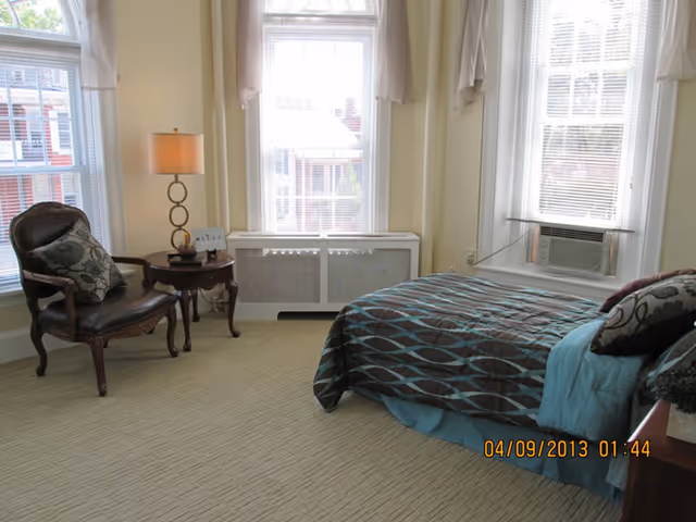 Sunny furnished bedroom with a bed, armchair, side table with lamp, and three tall windows.