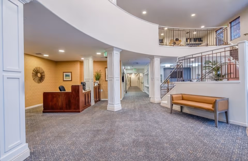 Interior view of a senior living facility lobby area with a wooden reception desk on the left, a long hallway in the center, a staircase with metal railing on the right, and a brown cushioned bench against the wall. The space has carpeted floors, white pillars, and warm lighting.