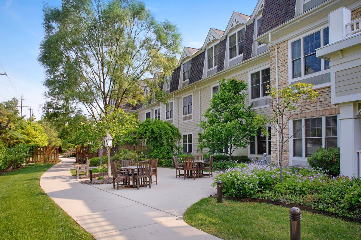Sunny courtyard with a paved walkway, wooden tables and chairs beside a multi-story residential building surrounded by trees and landscaping.