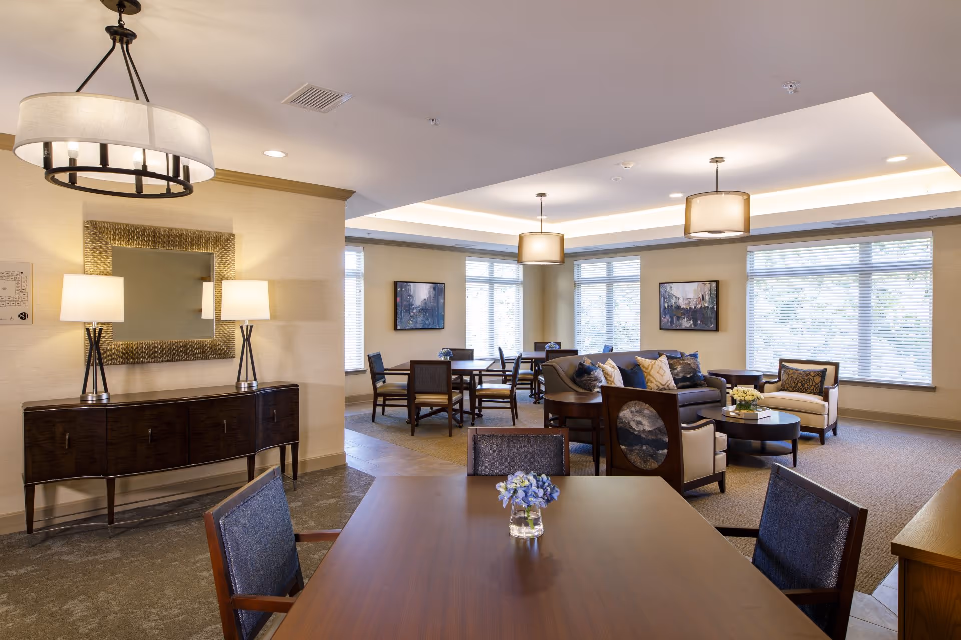 A well-lit common area in an assisted living facility featuring multiple seating arrangements including a wooden table with chairs and a small vase with flowers, a sofa with cushions, armchairs, and side tables. The room has large windows with blinds, two framed paintings on the walls, a decorative mirror above a dark wooden console table with two lamps, and modern ceiling light fixtures.