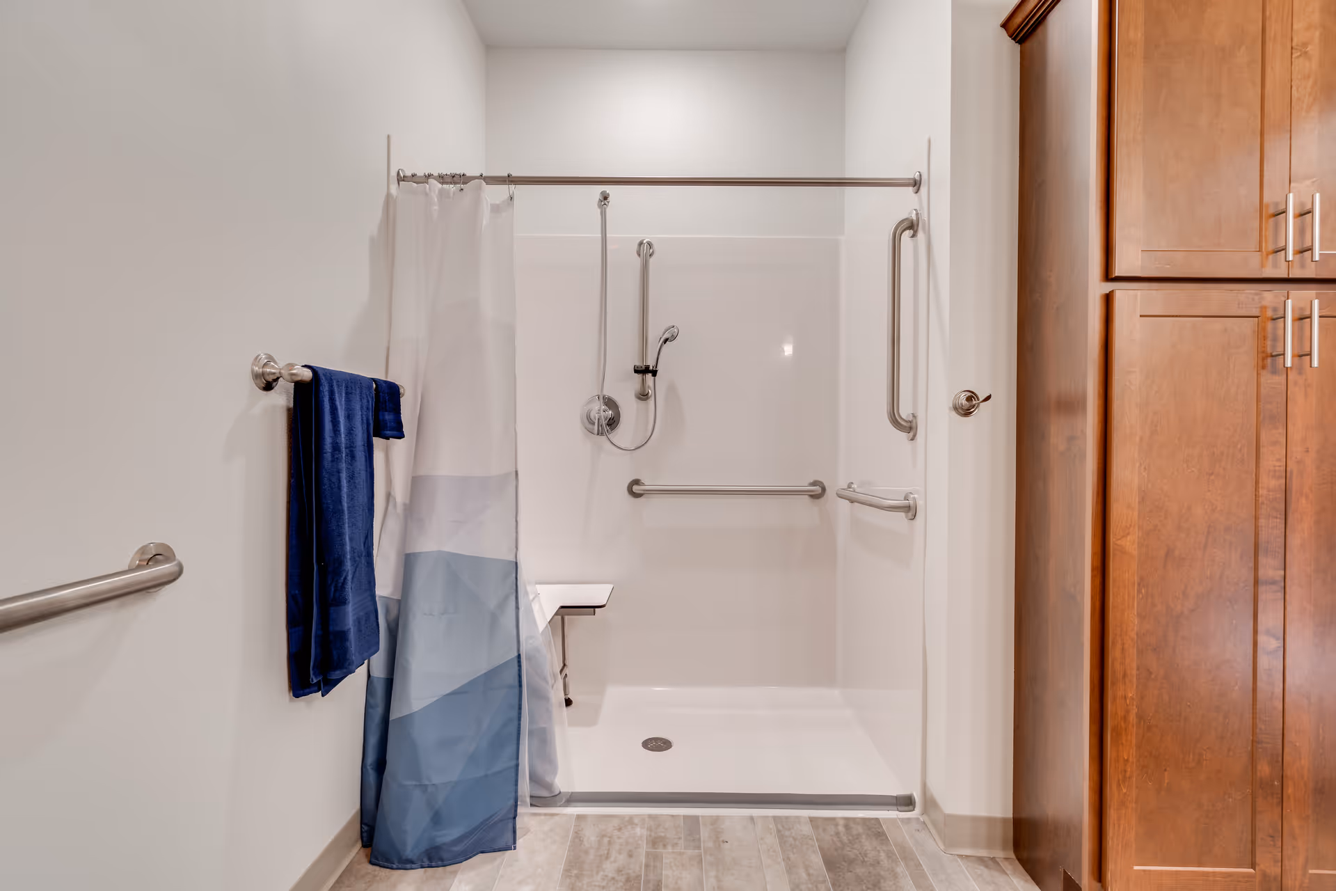 A clean, accessible shower area with a white and blue shower curtain, grab bars on the walls, a handheld showerhead, and a fold-down seat. There is a blue towel hanging on a towel rack to the left and wooden cabinets to the right.