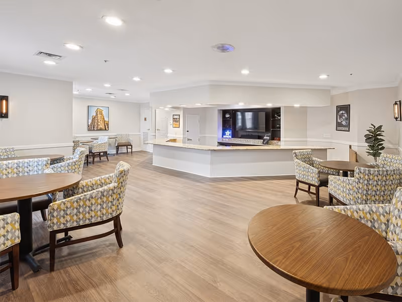 A bright and spacious common area with several round wooden tables surrounded by patterned upholstered chairs. The room features light-colored walls, wood flooring, recessed ceiling lights, and a large counter area with a TV mounted on the wall behind it. There are framed pictures on the walls and a potted plant in the corner.