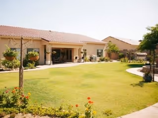 A spacious outdoor lawn area with green grass and flower beds in front of a single-story building with a tiled roof. The building has a covered patio with seating and potted plants, surrounded by trees and shrubs under a clear sky.