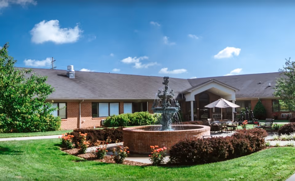 Front exterior of a single-story brick nursing facility with a central fountain, patio seating, and landscaped lawn under a blue sky.
