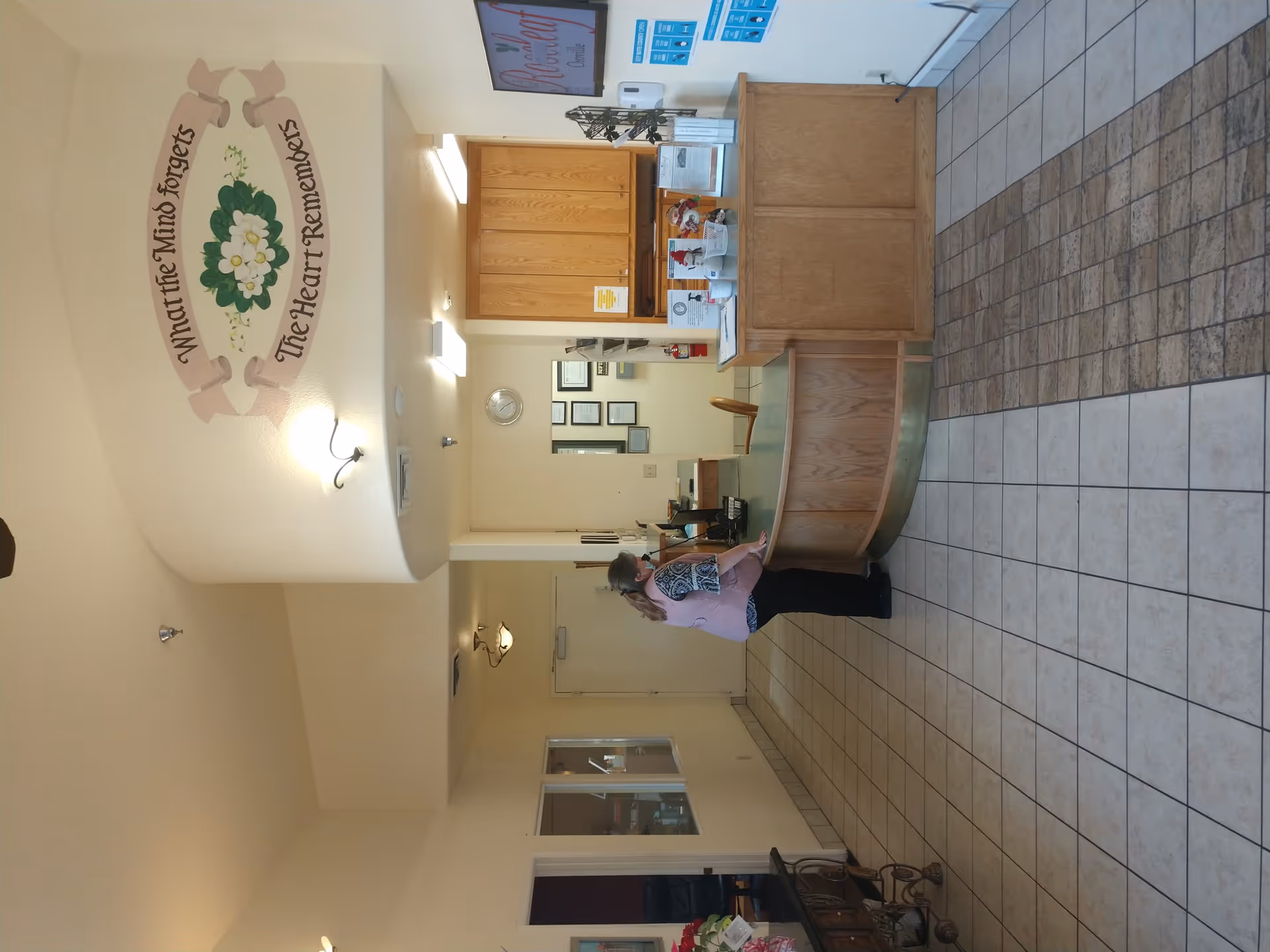Interior view of a reception area in a senior living facility with a curved wooden reception desk, a woman standing behind the desk, tiled floor, and a wall decoration with the quote 'What the Mind forgets The Heart Remembers'.