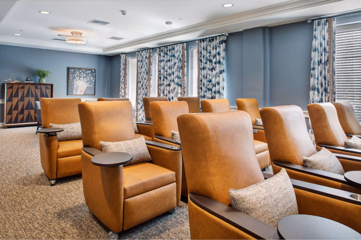 A cozy lounge area with multiple tan leather armchairs arranged in rows, each with a small attached round table and a decorative pillow. The room has blue walls, patterned curtains on the windows, a carpeted floor, and a wooden cabinet with a plant and artwork on the wall.