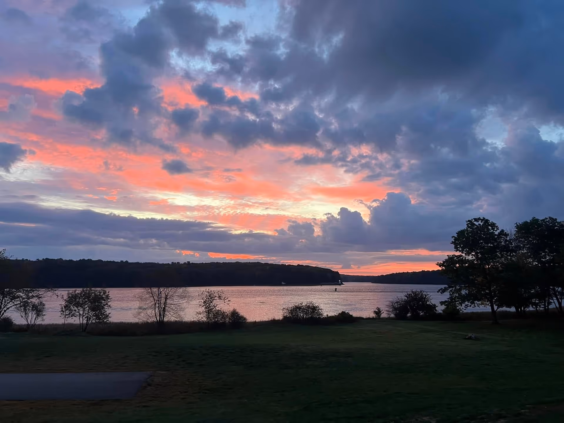 Scenic view of a lake at sunset with a partly cloudy sky showing pink and purple hues. Trees and grassy land are visible in the foreground and along the lake shore.