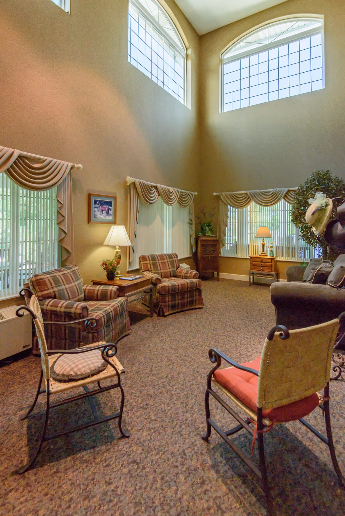 A cozy living room area with high ceilings and large arched windows letting in natural light. The room features two plaid armchairs, a wooden side table with a lamp and flowers, two metal-framed chairs with cushions, a dark sofa, and decorative window treatments. There are also plants and framed artwork on the walls.