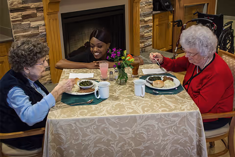 Two elderly women eating at a small dining table with plates and mugs while a smiling caregiver leans in by a fireplace.