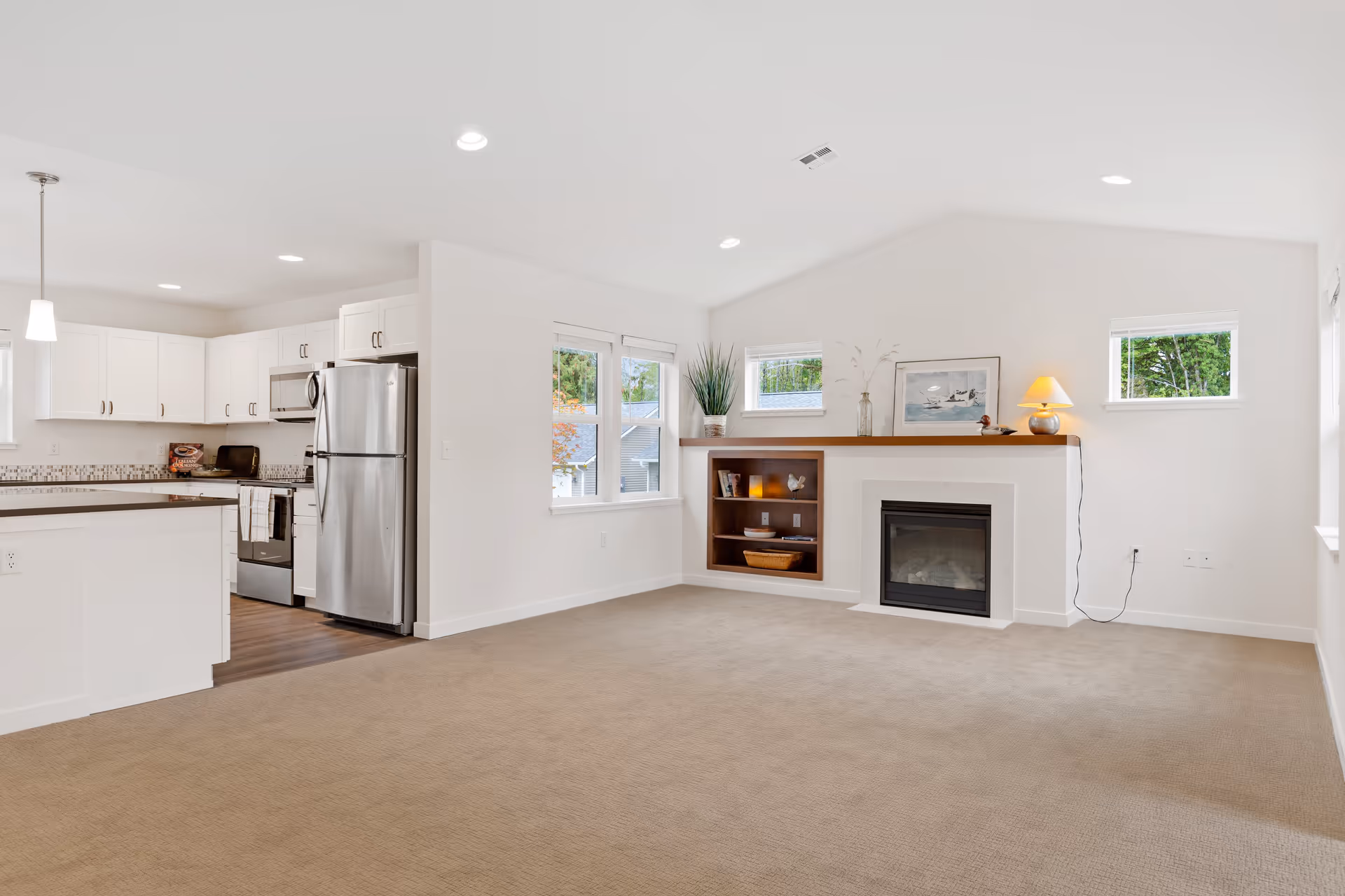Bright and spacious open-concept living area with beige carpet flooring, white walls, and recessed lighting. The room features a modern kitchen with white cabinetry, stainless steel appliances, and a breakfast bar on the left. On the right side, there is a cozy fireplace with a wooden mantel decorated with a lamp, framed artwork, and decorative items. Three windows provide natural light and a view of greenery outside.