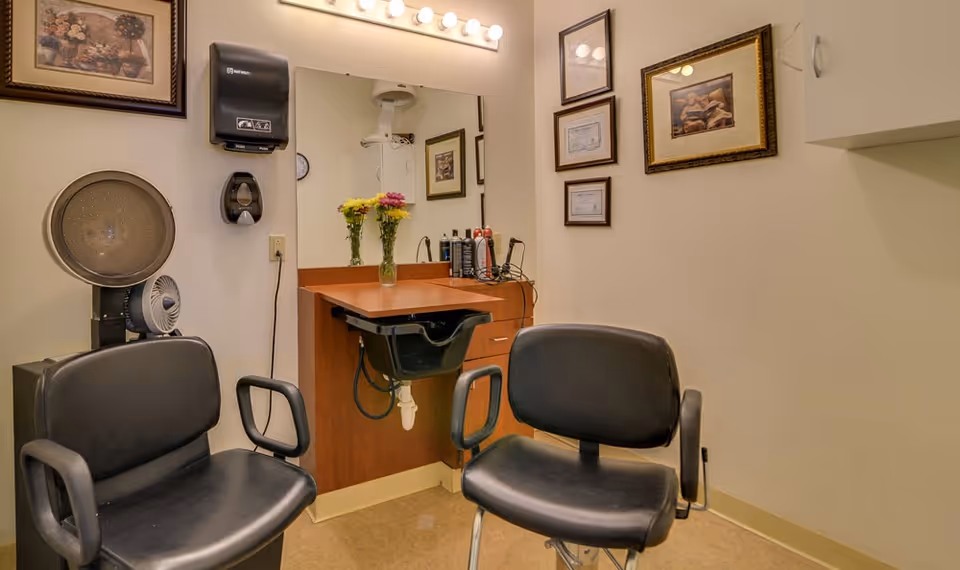 Interior view of a small hair salon area with two black salon chairs, a hair dryer, a large mirror with lights above it, a countertop with hair care products, and framed pictures on the wall.