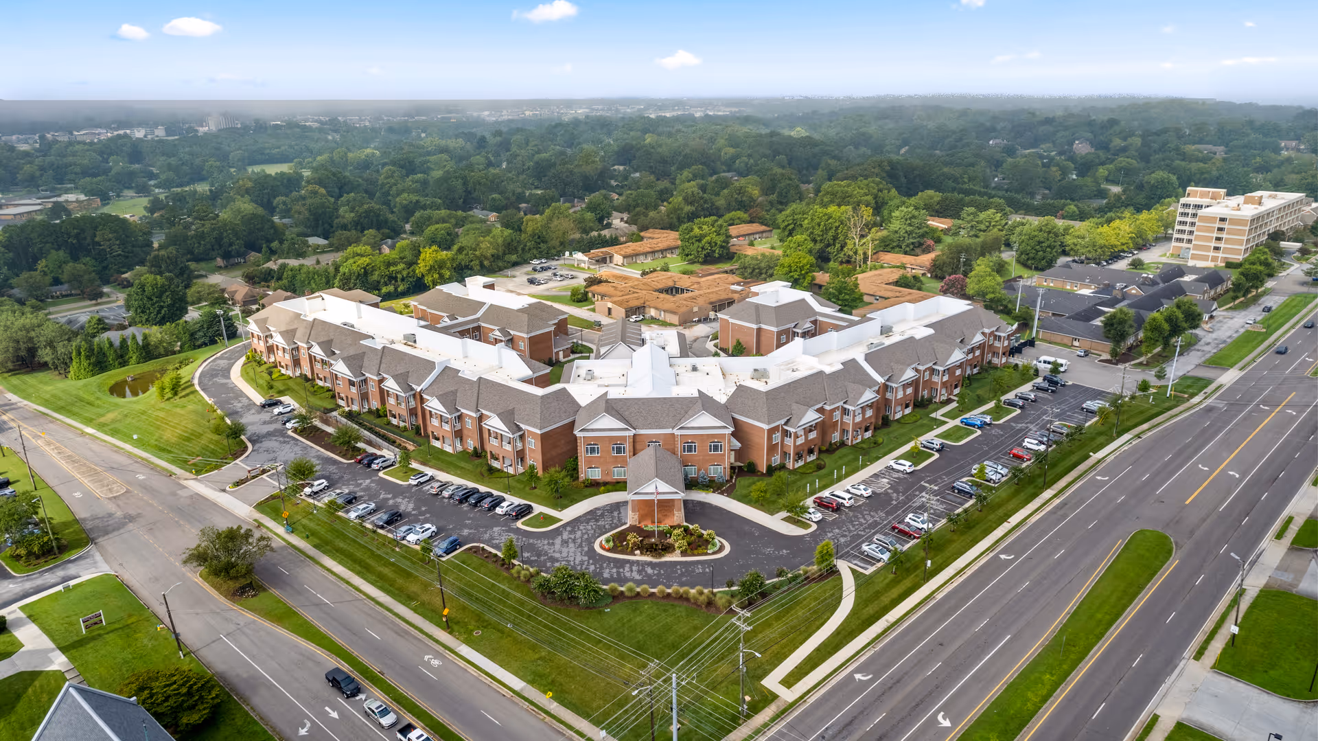 Aerial view of a large senior living facility with multiple connected brick buildings and white roofs, surrounded by parking lots and green lawns. The facility is located near a busy road with several cars parked in the parking areas. Trees and residential neighborhoods are visible in the background under a partly cloudy sky.
