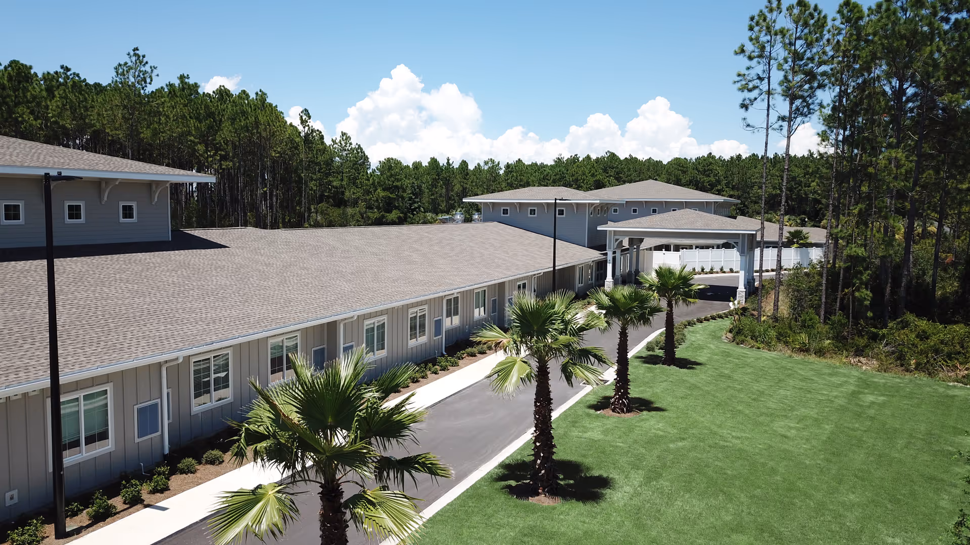 Exterior view of The Landing of Panama City Beach facility showing a long, single-story building with multiple windows, a driveway lined with palm trees, and a covered entrance. The building is surrounded by green grass and tall trees under a partly cloudy blue sky.