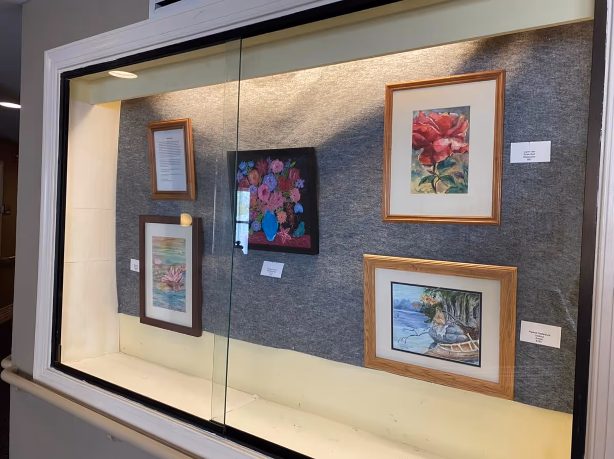 Glass display case in a hallway showcasing several framed paintings mounted on a gray felt background.