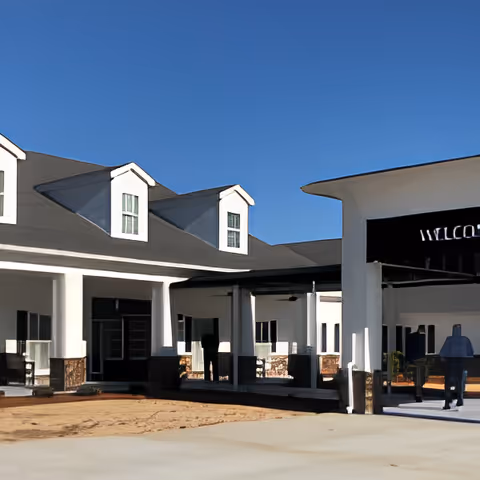 Exterior view of a senior living facility building with white walls, multiple windows, and a covered entrance area under a clear blue sky. A person is walking near the entrance.