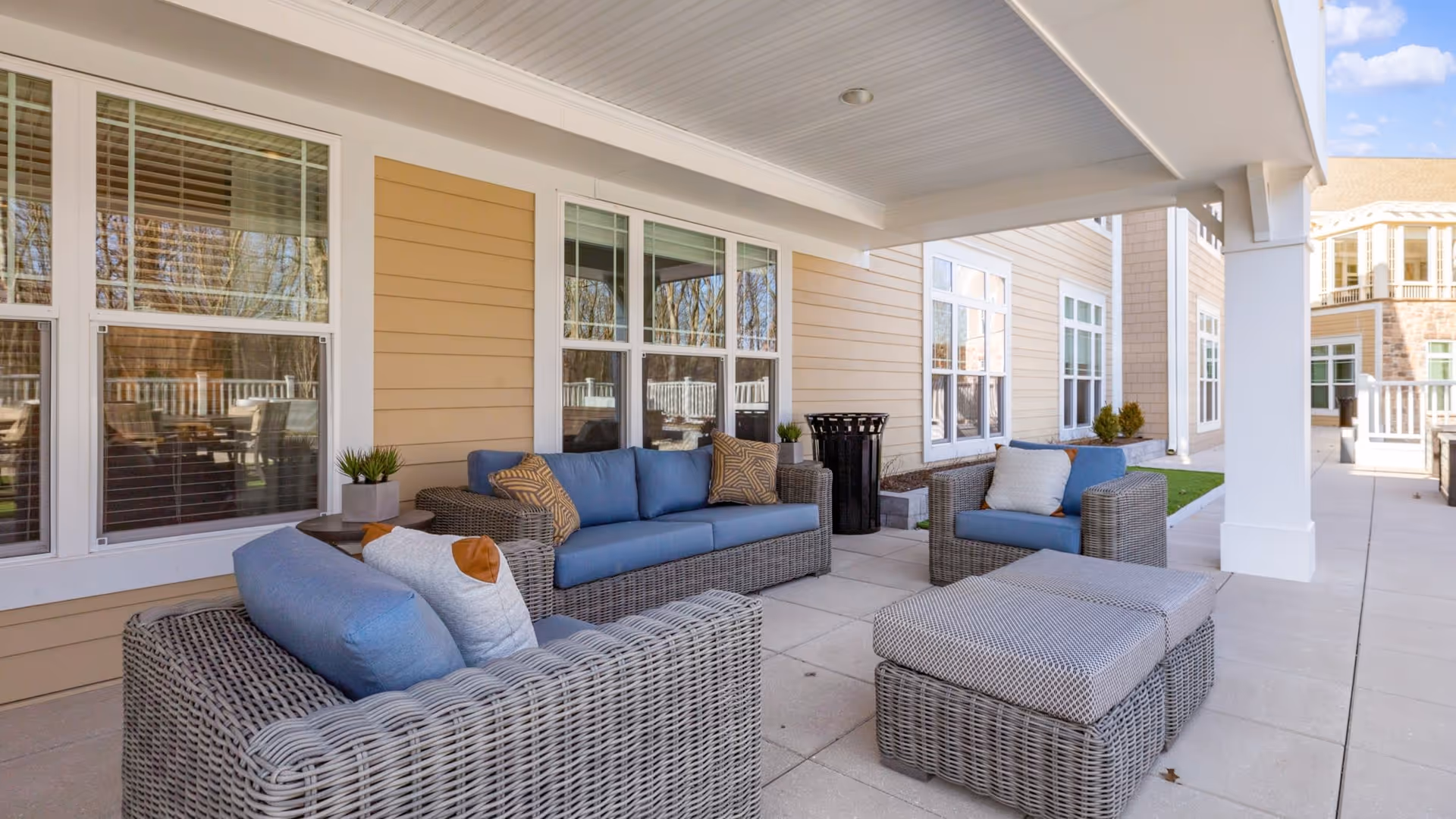 Covered outdoor patio area at Sunrise of Livingston with wicker furniture including two armchairs, a sofa with blue cushions and patterned pillows, and a matching ottoman. The patio is adjacent to a beige building with multiple windows and overlooks a paved walkway and landscaped area.