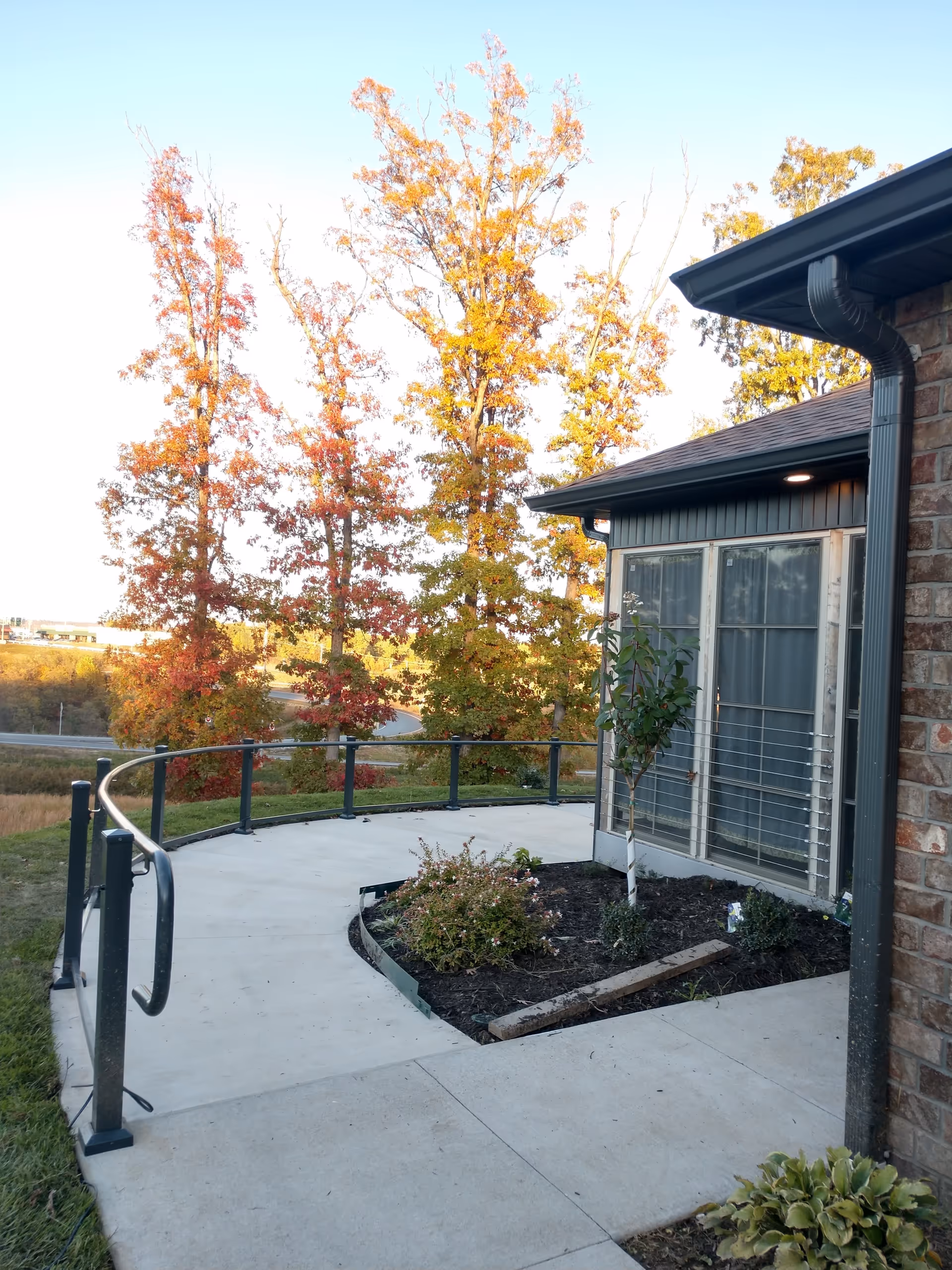 Curved concrete walkway with a black metal handrail leading to a building entrance with large windows. There is a small garden bed with plants and a young tree next to the walkway. In the background, there are tall trees with autumn-colored leaves under a clear sky.