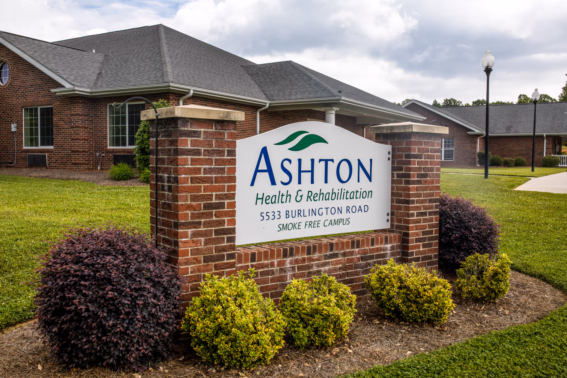 Outdoor view of Ashton Health & Rehabilitation facility sign made of white board with blue and green text, mounted on a brick structure surrounded by bushes and grass, with the building and lampposts visible in the background under a cloudy sky.
