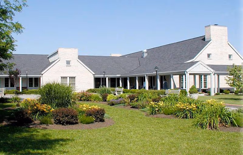 Exterior view of a single-story senior living facility building with a gray roof and white walls, surrounded by well-maintained green lawns and landscaped garden beds with shrubs and flowers under a clear blue sky.