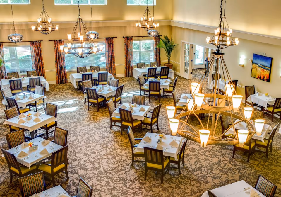A bright and spacious dining room with multiple tables covered in white tablecloths, each set with napkins and small flower arrangements. The room features large windows with colorful curtains, patterned carpet, and several modern chandeliers hanging from the ceiling. There is a potted plant near a doorway and a colorful painting on the wall.