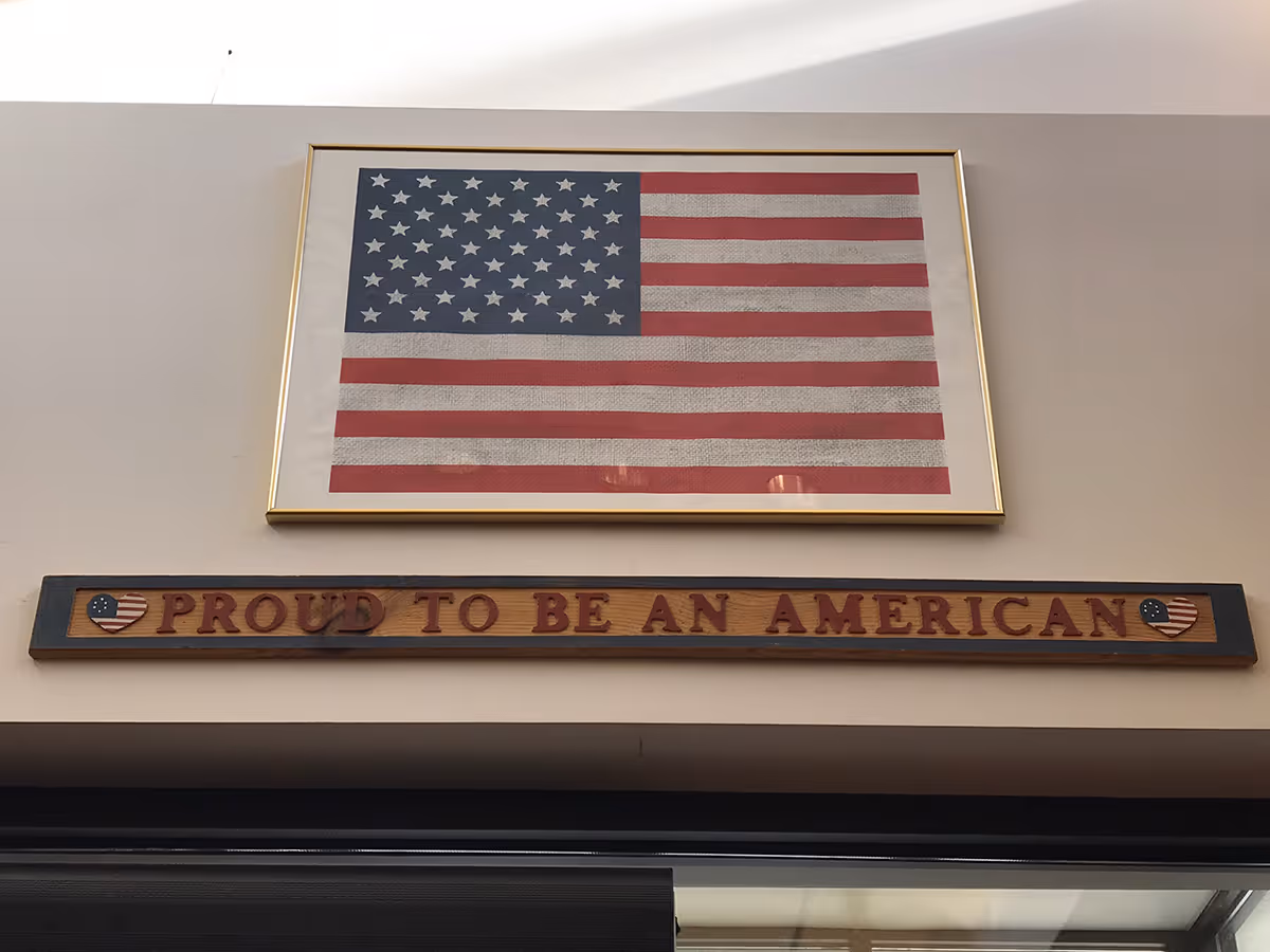 Framed American flag above a wooden sign that reads 'Proud to Be An American' mounted on an interior wall.