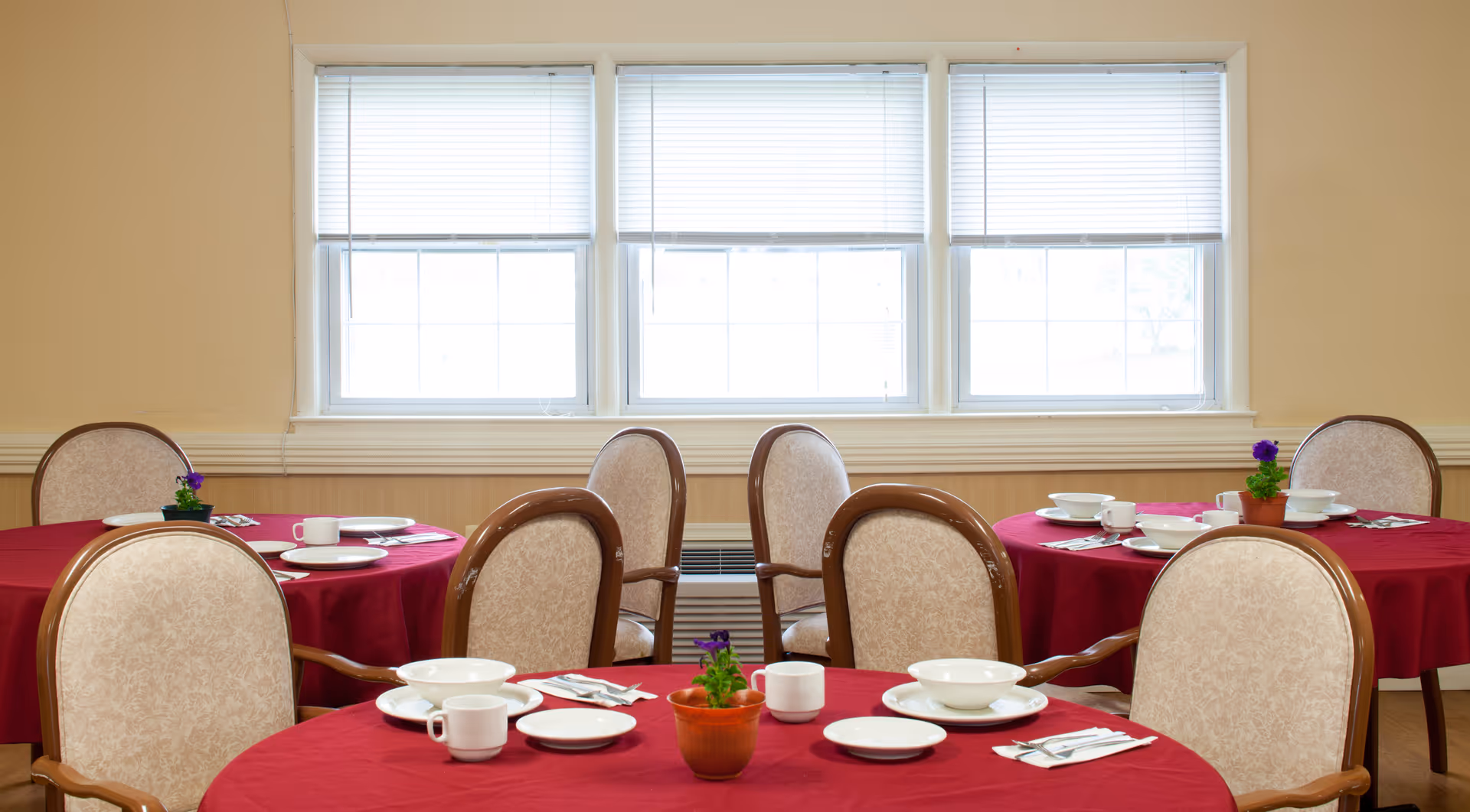 Dining room with round tables draped in red tablecloths, place settings and small potted plants, and upholstered chairs beneath three large windows.