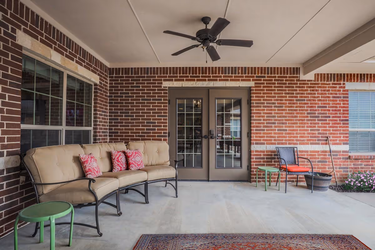 Covered outdoor patio area with a beige cushioned curved sofa adorned with three red patterned pillows, a small green side table, a black chair with an orange cushion, a potted plant, and double glass doors leading inside a brick building. A ceiling fan is mounted on the white ceiling above.