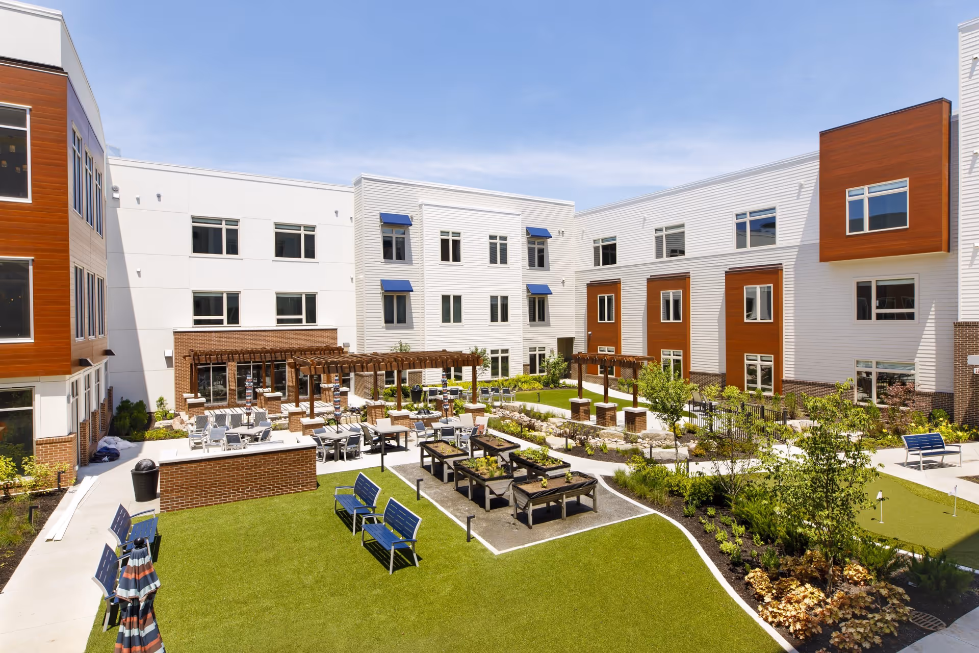Modern three-story assisted living building surrounding a landscaped courtyard with seating, raised garden beds, pergolas, and walking paths.