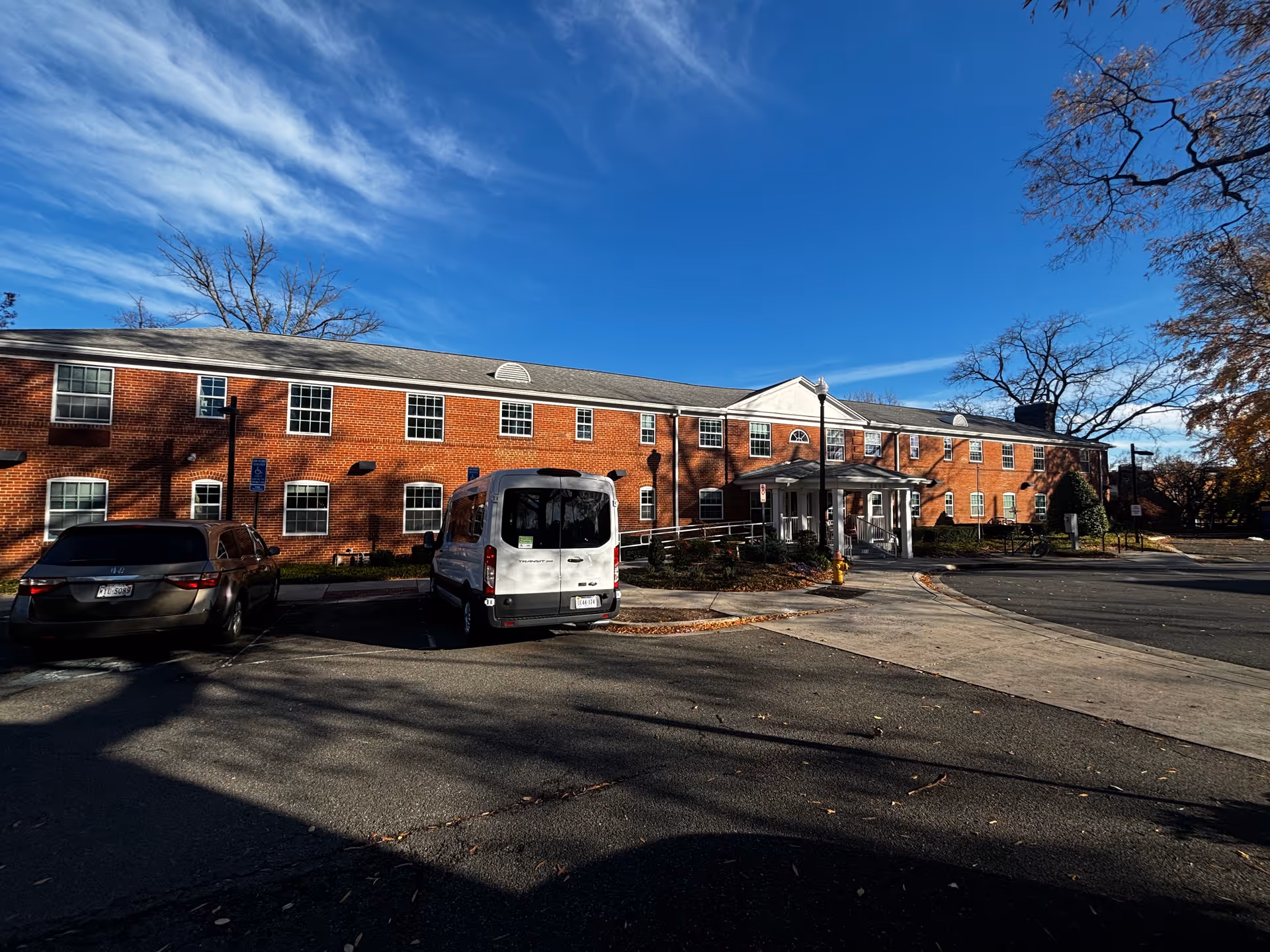 Exterior view of a two-story brick building with multiple windows and a covered entrance. There are two vehicles parked in front, a white van and a dark-colored car. The sky is clear with some wispy clouds, and there are leafless trees around the building.