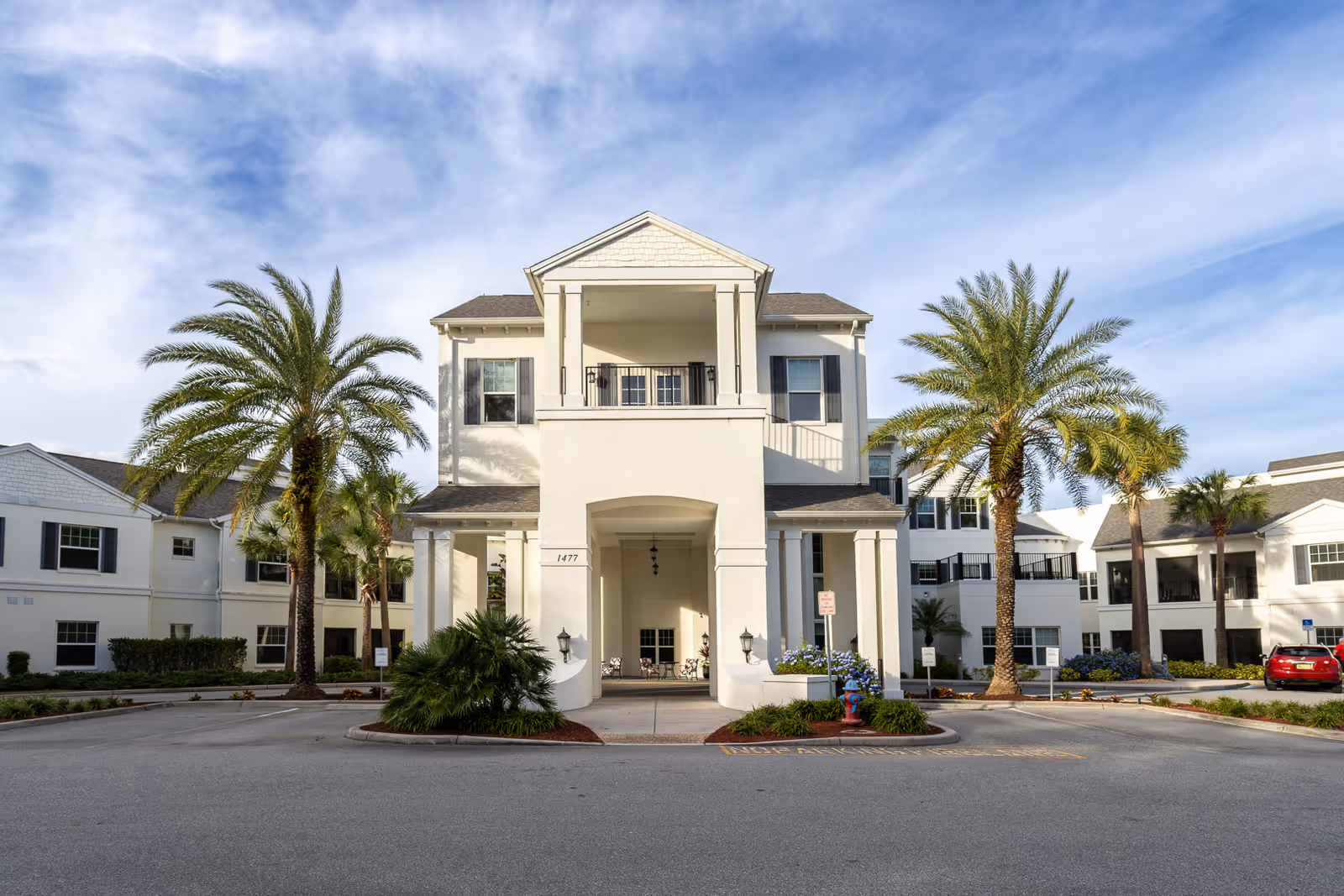 Front exterior view of a two-story white building with columns and a covered entrance, surrounded by palm trees and landscaping under a partly cloudy sky.