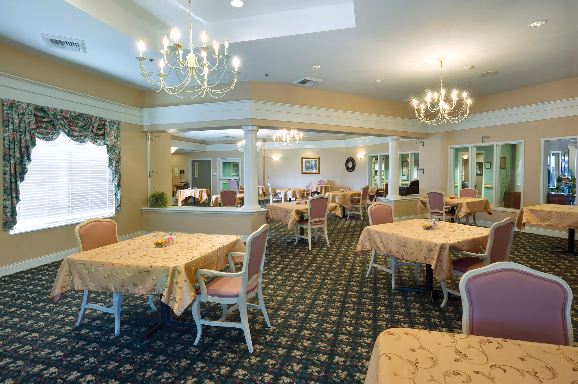 A spacious dining room with multiple tables covered in beige patterned tablecloths, each surrounded by pink cushioned chairs. The room features floral carpet, large windows with floral curtains, chandeliers hanging from the ceiling, and light-colored walls with decorative molding and columns.