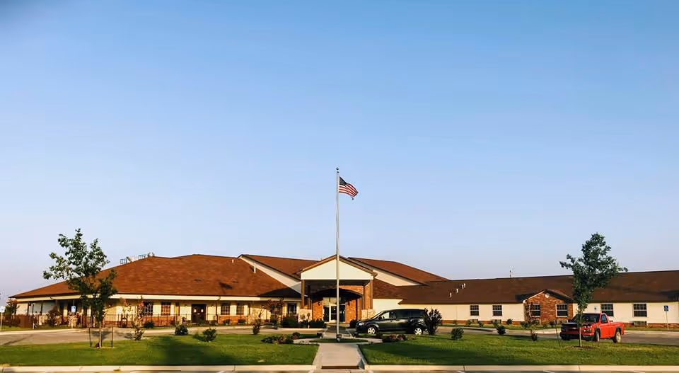 Front view of a single-story nursing and rehab building with a central entrance, an American flag on a flagpole, parked cars, and a lawn.