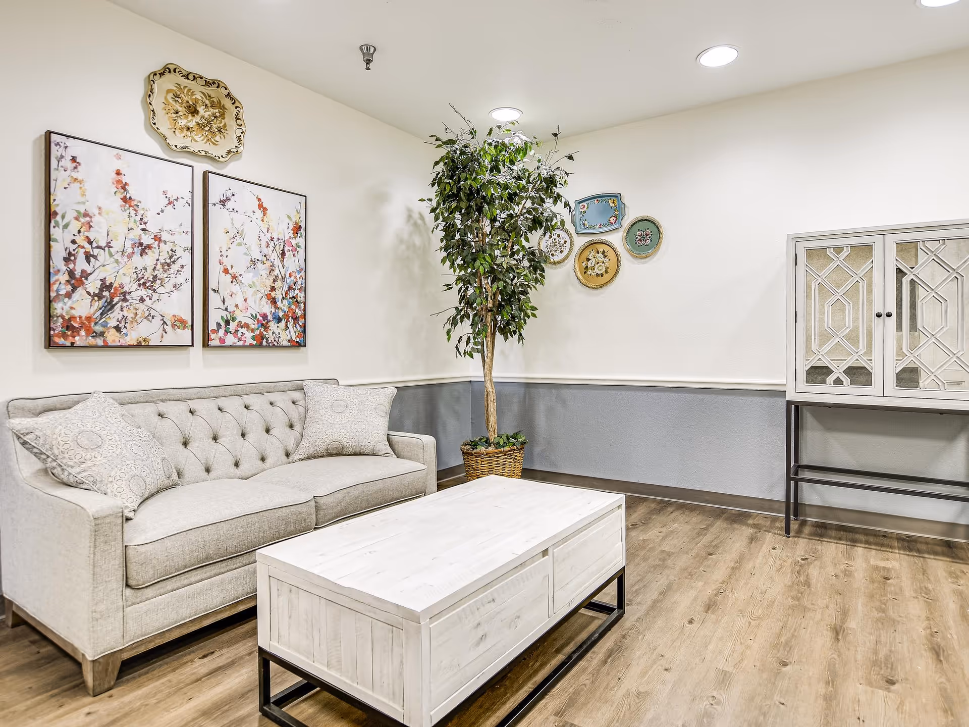 A cozy living room area with a light gray tufted sofa adorned with two patterned pillows, a white wooden coffee table with black metal legs, a tall potted artificial tree, and a decorative cabinet with mirrored doors. The walls are painted white with a gray wainscoting, and there are floral-themed framed artworks and decorative plates hanging on the walls.