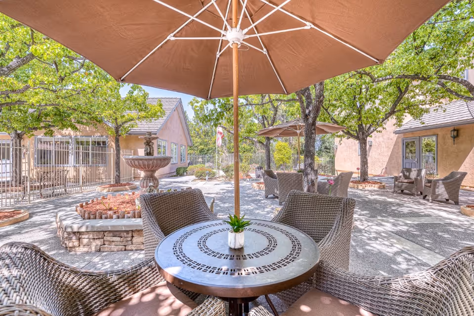 Outdoor patio area at Chino Hills Senior Living with round metal tables, wicker chairs, large brown umbrellas, trees providing shade, and a stone fountain surrounded by landscaping beds.