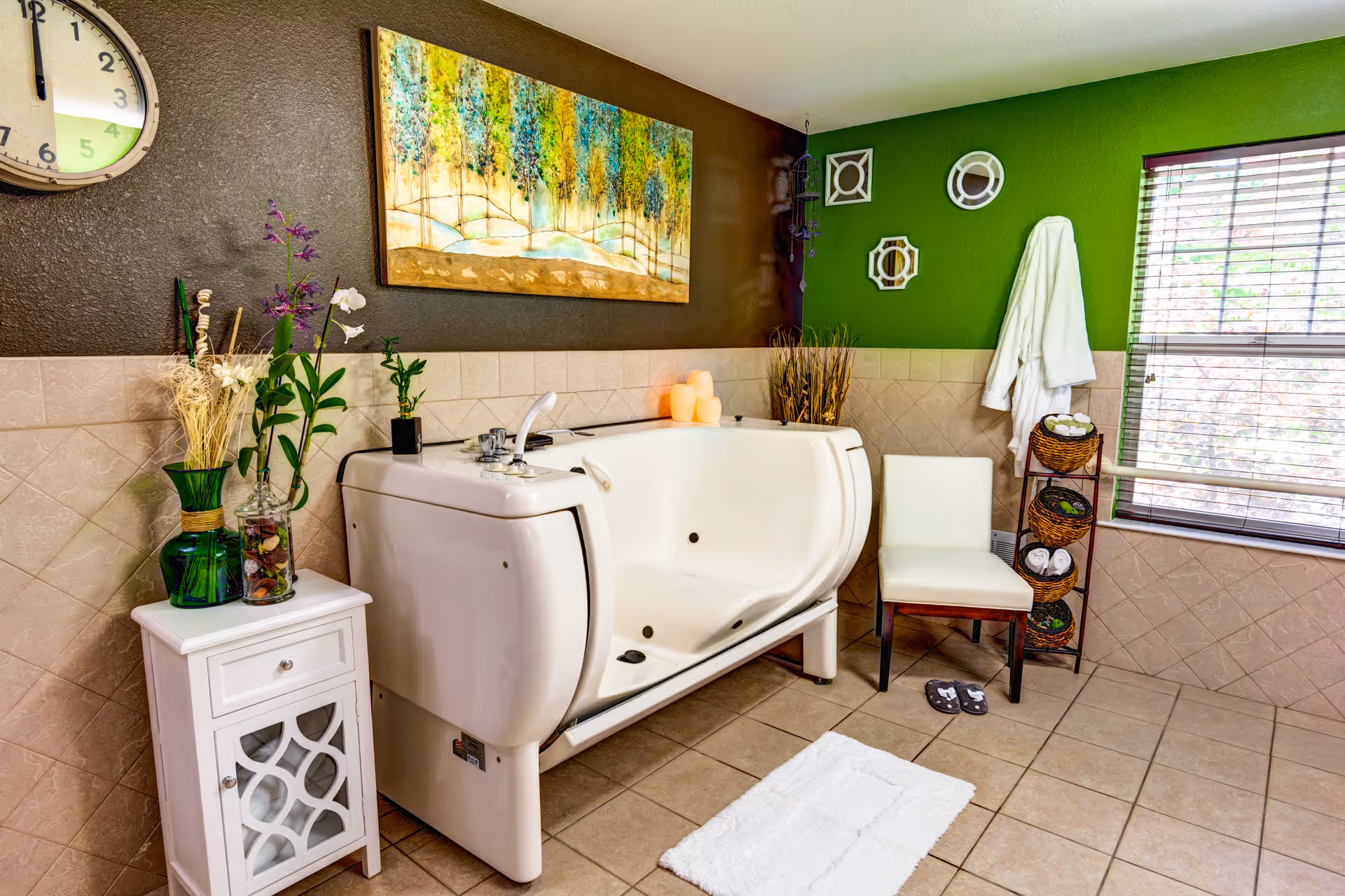 A bright bathroom featuring a large white walk-in bathtub with a faucet and controls. To the left of the tub is a small white cabinet with decorative vases and plants on top. Above the tub hangs a colorful painting of trees. On the right side, there is a white chair next to a rack holding rolled towels and a white bathrobe hanging on the green wall. A window with blinds allows natural light to enter the room. The floor is tiled, and a white bath mat is placed in front of the tub.