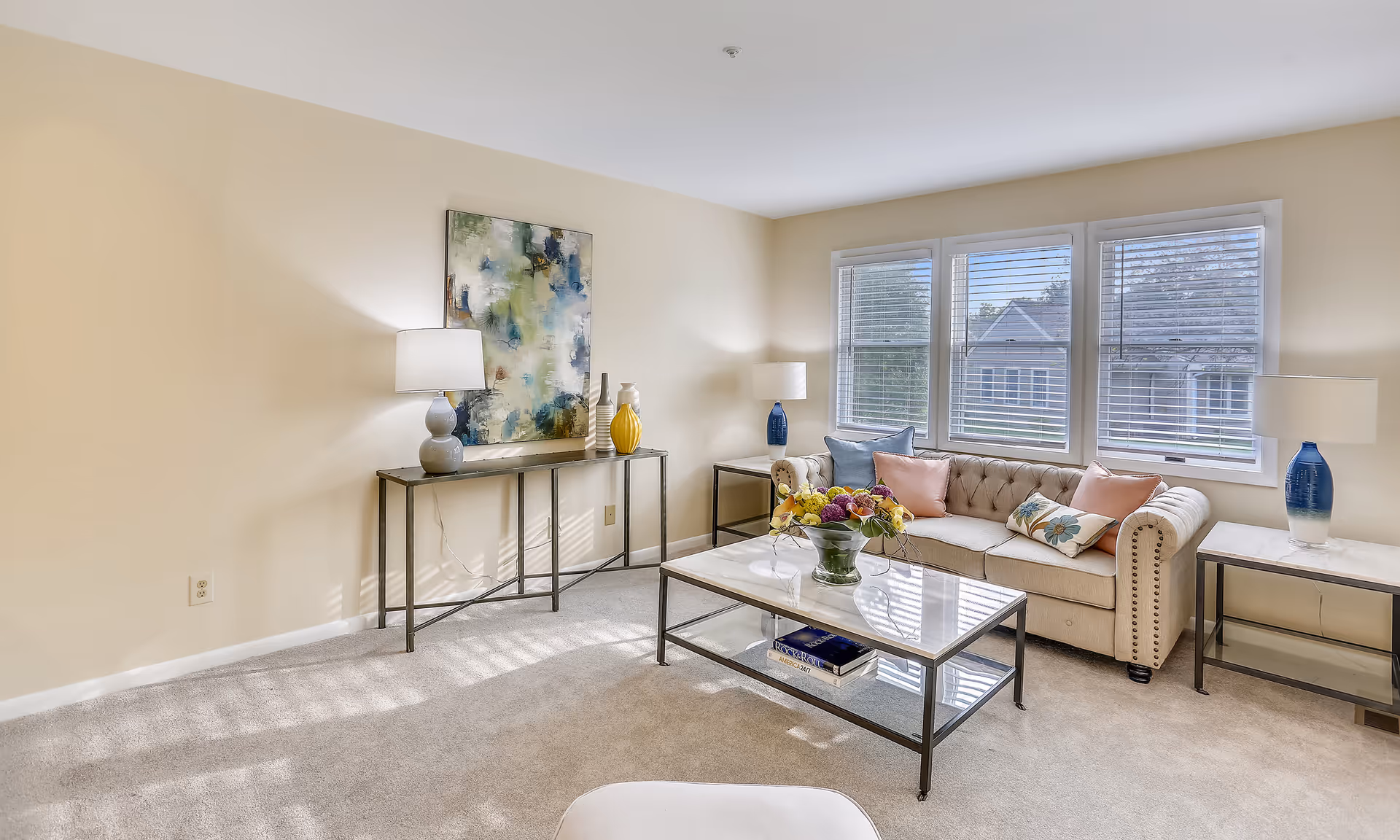 A bright living room with beige walls and carpet, featuring a beige tufted sofa with colorful pillows, a glass coffee table with a flower arrangement, two side tables with blue and white lamps, and a console table with decorative vases and a large abstract painting above it. Three large windows with white blinds allow natural light to fill the room.