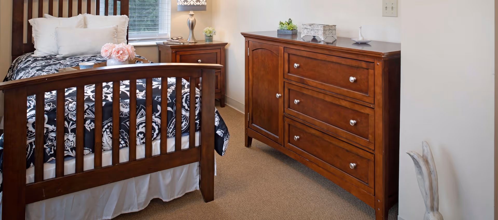 Bedroom with a wooden slatted bed and matching dresser, black-and-white patterned bedding, nightstand and lamp.