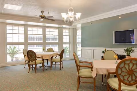 Bright dining area with round tables, upholstered chairs, large windows, and wall-mounted TV.